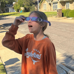 Girl in orange sweatshirt wearing solar eclipse glasses